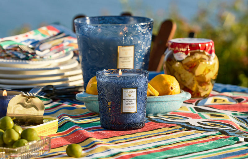 Voluspa Mediterranean Lemon candle styled on a colorful outdoor table with fresh lemons and grapes