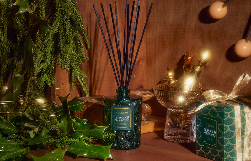 Voluspa Noble Fir Garland reed diffuser styled with fresh evergreen garland and holly against a dark wood backdrop