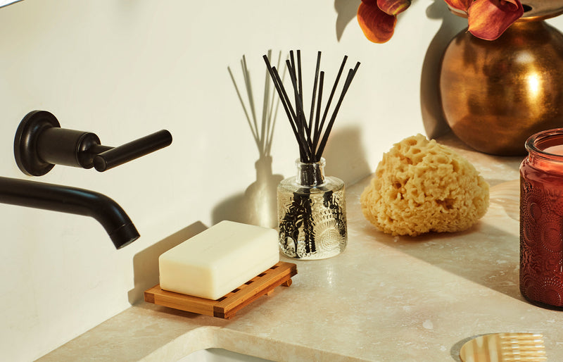 Reed diffuser with black reeds on a bathroom vanity beside soap and a natural sponge